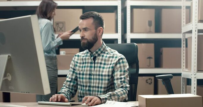 Logistics manager sits at desk in warehouse on background of shelves with cardboard boxes and works with documentation on computer.
