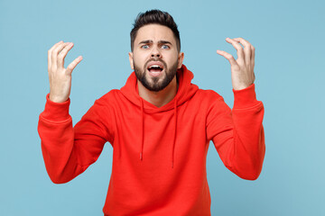 Young caucasian angry indignant nervous irritated stressed bearded man 20s wearing casual red orange hoodie spreading hands isolated on blue color background studio portrait People lifestyle concept.