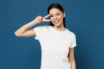 Young smiling pretty happy cute cheerful positive attractive latin woman 20s wear white casual basic t-shirt show covering eye with victory gesture isolated on dark blue background studio portrait.