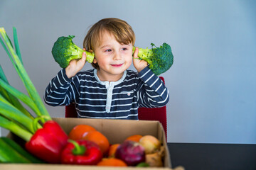 Fototapeta premium Little boy sitting at the table and taking broccoli in his hand. Set of different fresh vegetables in a cardboard box. Teaching child to healthy and varied vegitarian food.