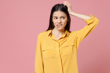 Young overthinking worried thoughtful pensive troubled brunette attractive latin woman in yellow shirt sctratch back head looking aside oops gesture isolated on pastel pink background studio portrait.