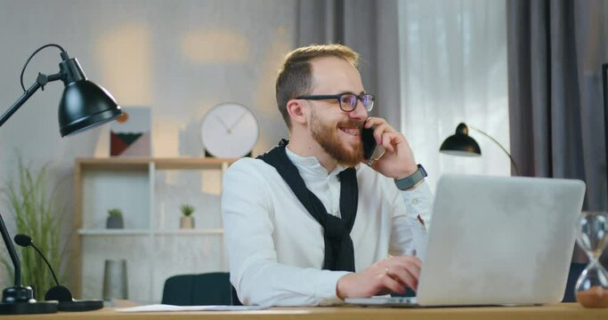 Handsome cheerful young bearded guy in glasses laughing from joke that hearing from his interlocutor on phone during working on laptop at home
