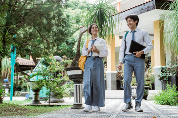 couple of Indonesian high school students smiling while walking carrying book and laptop with the trees and school building background