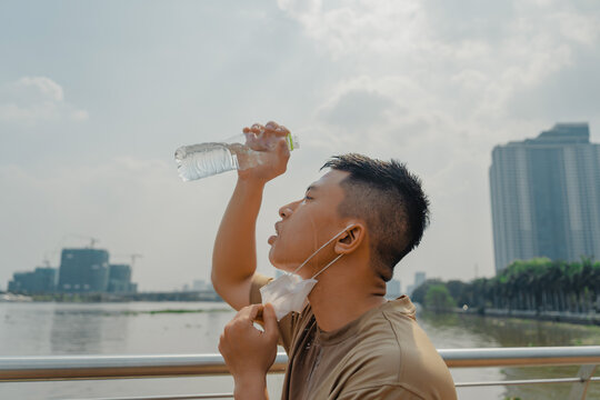 Young asian runner man putting off face mask while using water in bottle to wash his face after finished running and working out.