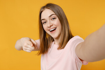 Close up young leader woman 20s with nude make up wearing casual basic pastel pink t-shirt doing selfie shot on mobile phone point finger camera on you isolated on yellow background studio portrait