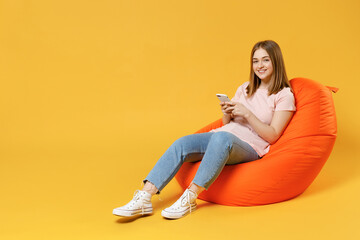 Full length of young woman 20s in basic pastel pink t-shirt, jeans sitting in orange bean bag chair holding mobile cell phone chatting browsing internet isolated on yellow background studio portrait.