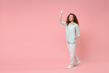 Fulll length young black african american happy smiling friendly curly woman 20s in blue shirt looking camera walk going waving hand greet someone isolated on pastel pink background studio portrait.