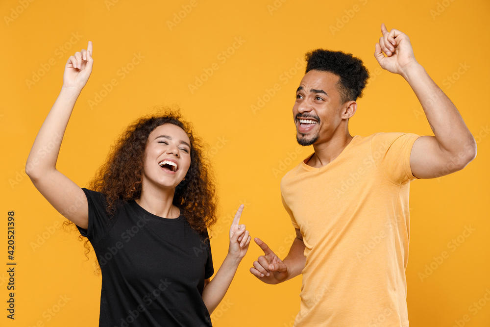 Wall mural Young couple two happy friends family african overjoyed excited man woman 20s wearing black t-shirt do winner gesture point index finger up celebrating isolated on yellow background studio portrait.