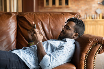 Cozy rest. Peaceful millennial black guy hipster lying on back on couch spend leisure time in internet using phone app. Calm relaxed young african man enjoy serene weekend alone read news chat online