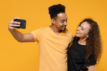 Young couple two friends together family african smiling happy man woman 20s wear black t-shirt do selfie shot on mobile phone look to each other isolated on yellow color background studio portrait.