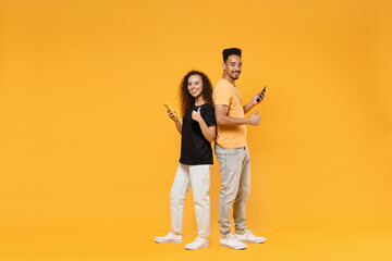 Full length young couple two friends fun together african happy woman man in black tshirt show hold mobile phone stand back to back show thumb up gesture isolated on yellow background studio portrait