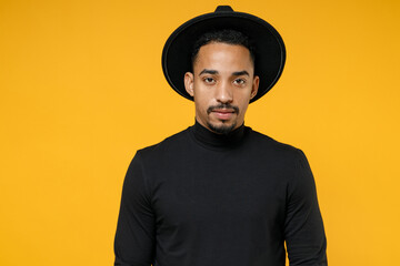 Young african american serious fashionable attractive man 20s wearing trendy stylish black hat shirt looking camera isolated on yellow orange color background studio portrait People lifestyle concept