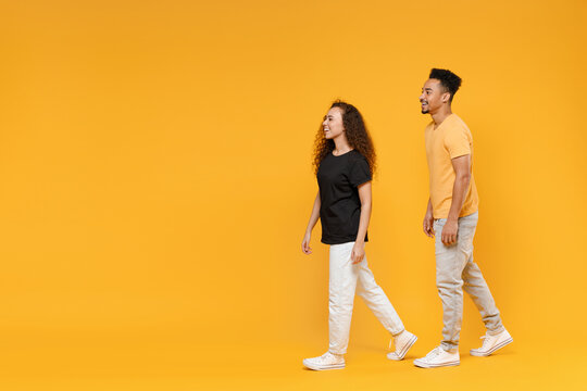 Full length side view young couple together two fun friends family happy fun smiling african man woman 20s in yellow black t-shirt walking going isolated on orange color background studio portrait.
