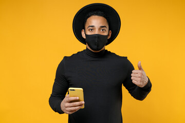 Young african man wear stylish black hat shirt face mask to safe from coronavirus covid-19 pandemic quarantine hold mobile phone show thumb up gesture isolated on yellow background studio portrait.