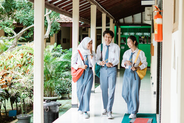 Three high school students wearing school bags and carrying a smartphone while walking together in the corridors of the school building