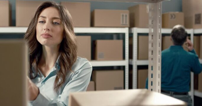 Female logistics manager prepares goods in warehouse for export and scans barcodes on cardboard boxes entering data into tablet.