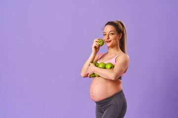 attractive, happy pregnant woman holds fresh organic green apples while standing on purple background. expectant mother eats an apple. Healthy, proper nutrition. Vegetarian, dietary food. Vitamins.