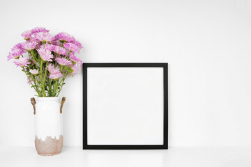 Mock up square black frame with vase of purple flowers. White shelf against a white wall. Copy space.