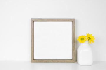 Mock up square wood frame with vase of yellow daisy flowers. White shelf against a white wall. Copy...