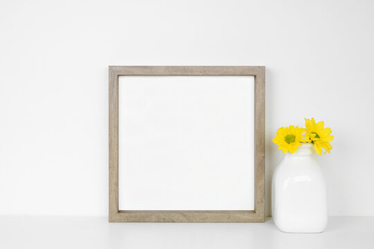 Mock up square wood frame with vase of yellow daisy flowers. White shelf against a white wall. Copy space.