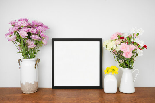 Mock up square black frame with vases of colorful flowers. Wooden shelf against a white wall. Copy space.
