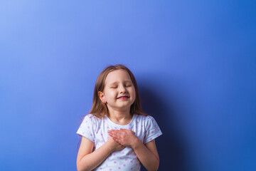 little girl with her eyes closed holds her hands on chest, feels grateful, child dreams with friendly expression, pressing her palms to her chest. concept of love and faith on a purple background