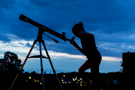 Young Girl Looking through Telescope at Moody Sunset