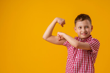 positive, confident Caucasian 8-year-old in shirt smiles and raises his clenched fists, flexing muscles in his arms, feeling strong and full energy after eating healthy protein lunch and working out