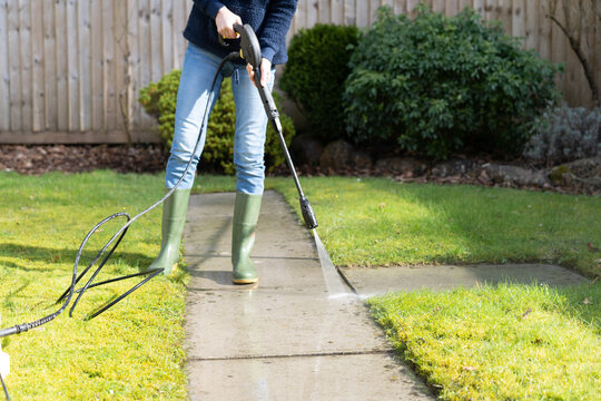 Woman In Waterproof Boots Power Cleaning The Garden Back Yard Paving Slabs  Using A High Pressure Hose With Jet Attachment