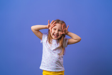 Fototapeta na wymiar little girl 5 years old with blonde hair in a T-shirt with colored polka dots stands smiling and holding her hands to her face on purple background. model is having fun and feels overwhelmed, amazed.