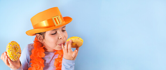 King's Day in Holland. Traditional festival on April 27 in the Netherlands. A little girl in a festive orange hat on a blue background eats colored donuts. Banner, long format. Copy space