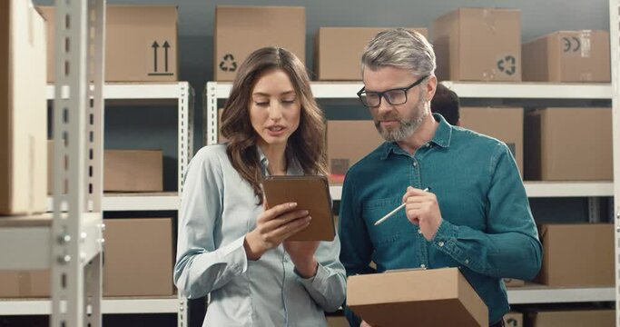 Two colleagues, logistics manager, man and woman, stand in the middle of warehouse and inspect parcel, recording information on tablet.