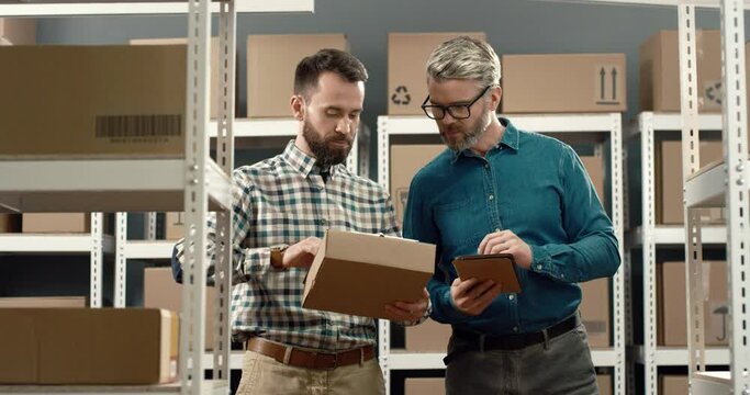 Two male managers stand in warehouse with cardboard boxes and solve problems with indefinite parcels. Logistics work.