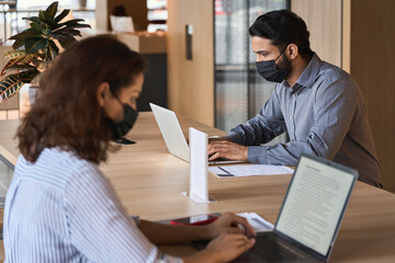 Indian business man student wearing face mask working on laptop safe distancing. Diverse people in facemasks using computers sitting at table with social distance sign in office coworking or cafeteria