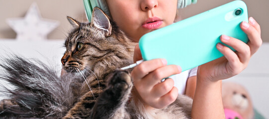 Child with headphones playing a computer game on the phone. A little girl lies on the bed and fluffy cat a looks smartphone. The concept of hobbies and hobbies of modern teenagers. Banner
