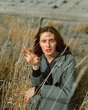 Young Woman In Coat Sitting In Dry Grass And Looking Forward