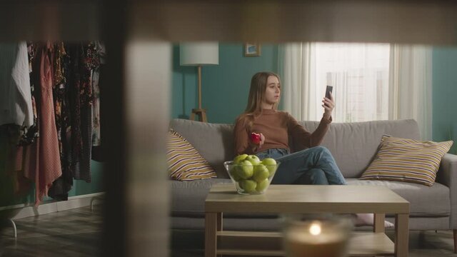 Pretty girl in blue jeans and brown sweater sits on sofa on window background, bites red apple and talks on video call. On coffee table is glass jar with green apples. Camera dollies horizontally