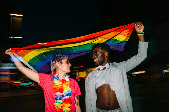 Happy multiracial couple raising LGBT pride flag in street at ni