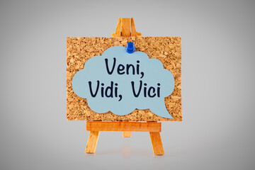 Blue speech bubble with Latin phrase Veni, vidi, vici on corkboard on wooden easel.