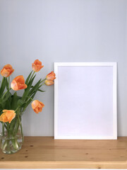 A white empty frame on the table for a photo or painting in the interior and delicate orange-pink tulips in a glass vase next to it