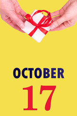 October 17th. Festive Vertical Calendar With Hands Holding White Gift Box With Red Ribbon And Calendar Date 17 October On Illuminating Yellow Background. Autumn month, day of the year concept.