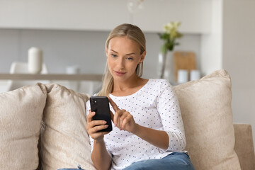 Young Caucasian woman relax on sofa in living room look at cellphone screen text message online. Millennial female talk speak on video call on smartphone or shop on web on cell. Virtual event concept.