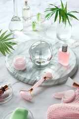 Obraz na płótnie Canvas Gua Sha massager and pink quarz face roller on marble stone background. Face care products, glass crystal ball and exotic palm leaves. Towel, skincare tonic, essential oil and revitalizing serum.