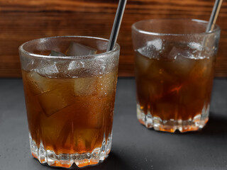 Iced coffee in a low glass on a black background with a wooden backdrop. Cold summer drink. Selective focus on the front wall of the front glass. Water drops on a glass. Cocktail tubes
