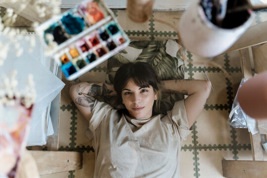 Top view of happy young woman lying on floor with watercolor and paint on glass table.