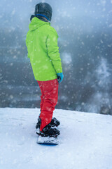 Fototapeta na wymiar Snowboarder in bright sports green jacket standing on his board on top of slope. Winter outdoor activities