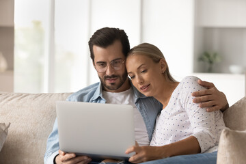 Fototapeta premium Happy young Caucasian couple relax on sofa at home look at laptop screen watch video online together. Millennial man and woman rest on couch use computer browsing internet or shopping online.