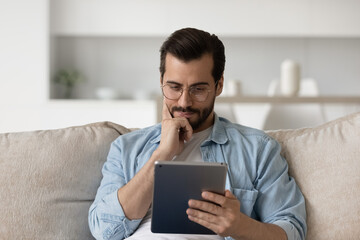 Pensive millennial Caucasian man relax on sofa at home use tablet thinking pondering. Unhappy...