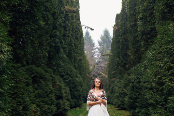 bride stands among trees in park in wedding dress and a shawl on
