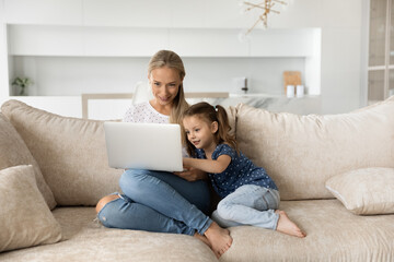 Smiling young Caucasian mom and small daughter sit on sofa at home use modern laptop together. Happy mother and little girl child relax on couch study online talk on video call on computer.